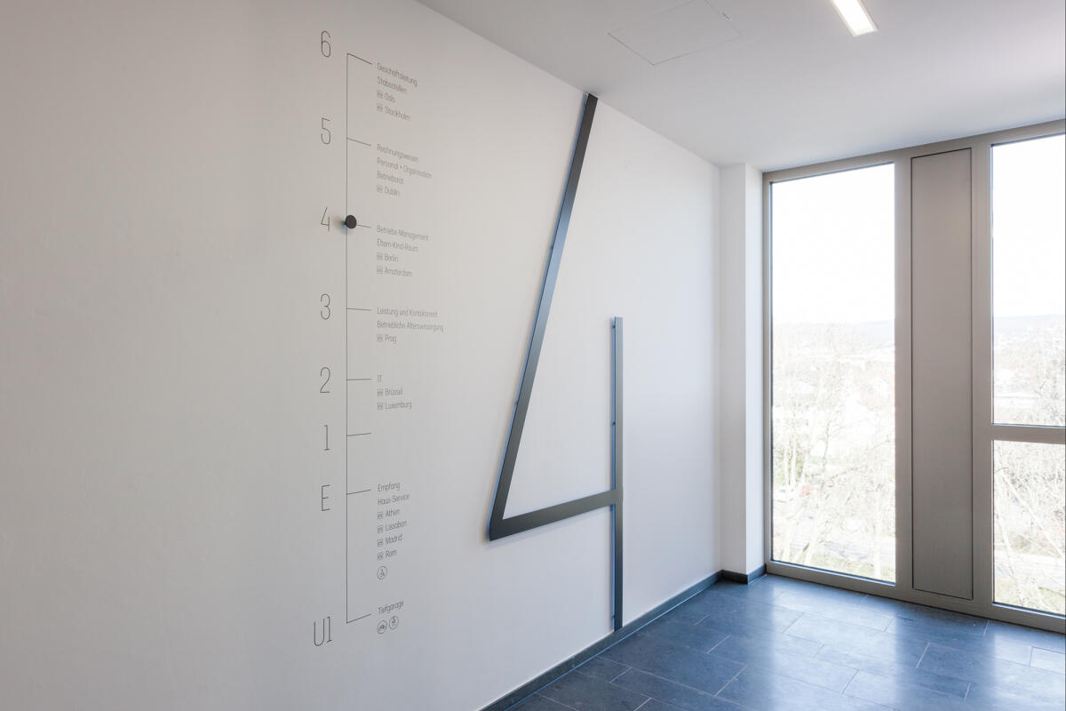 An der Wand befindet sich eine lineare Grafikanzeige, die Maße in einer vertikalen Anordnung darstellt. Neben dieser Anzeige ist ein großer, schräg verlaufender Metallstreifen angebracht, der eine dynamische Form bildet. Auf der gegenüberliegenden Seite des Raumes sind große Fenster, die Licht hineinlassen und einen Ausblick auf die Umgebung bieten.