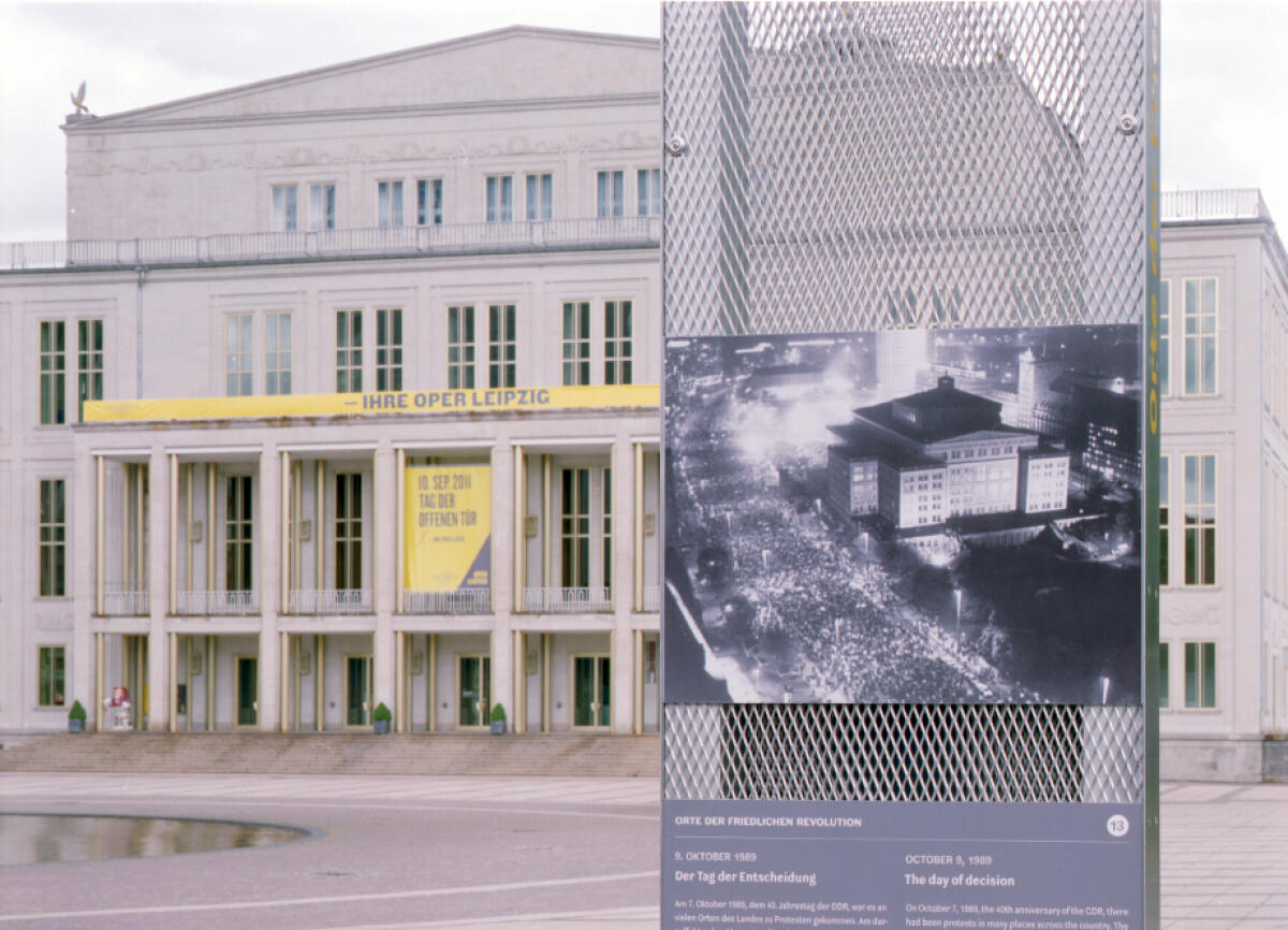Das Bild zeigt die Fassade der Oper in Leipzig, ein großes, monumentales Gebäude mit hellen Wänden und mehreren Fenstern. Im Vordergrund steht ein Informationstafel mit einer historischen Fotografie von einer Menschenmenge vor einem anderen Gebäude, die in der Nacht aufgenommen wurde. Darüber hängt ein gelbes Banner mit dem Schriftzug 'Die Oper Leipzig'.