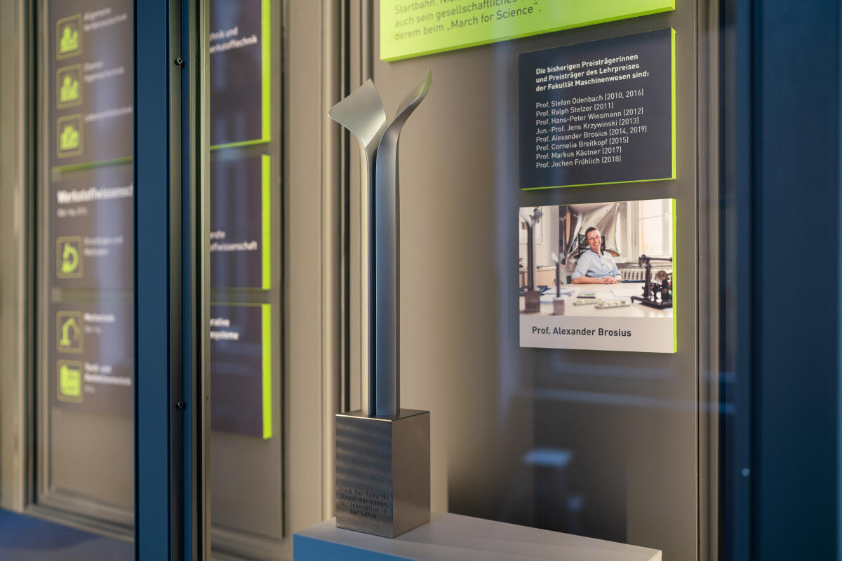 Das Bild zeigt eine Vitrine mit einem silbernen, modernen Preis oder einer Auszeichnung, die auf einem Podest steht. Im Hintergrund befinden sich Informationsbanner mit grünen Akzenten, die Namen und Details zu verschiedenen Personen oder Präsentationen enthalten. Neben der Vitrine ist ein Foto von einer Person, die in einem Büro sitzt und an einem Tisch arbeitet.