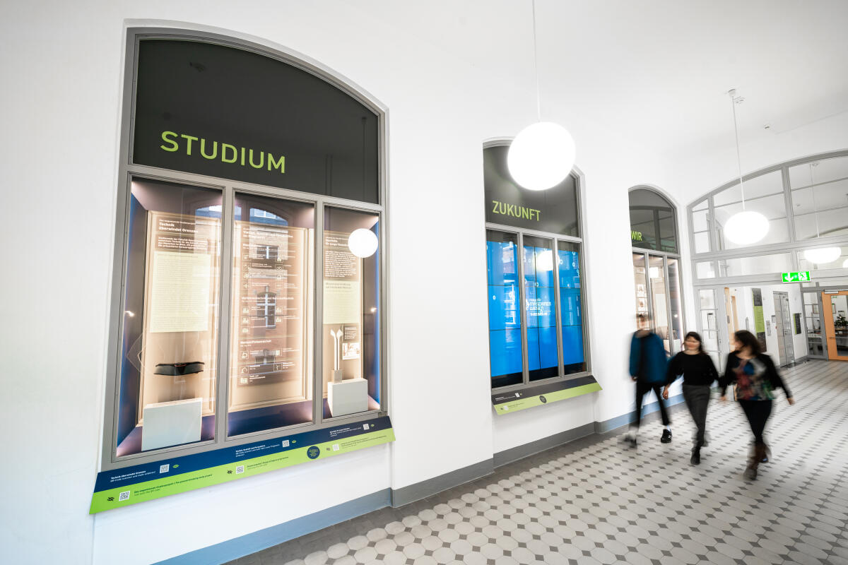Das Bild zeigt einen hellen Flur mit drei Fenstern, die Informationen präsentieren. Links steht 'STUDIUM' und rechts 'ZUKUNFT', beide mit Beschriftungen und beleuchtet. Im Vordergrund sind verschwommene Silhouetten von Personen, die den Flur entlanggehen.