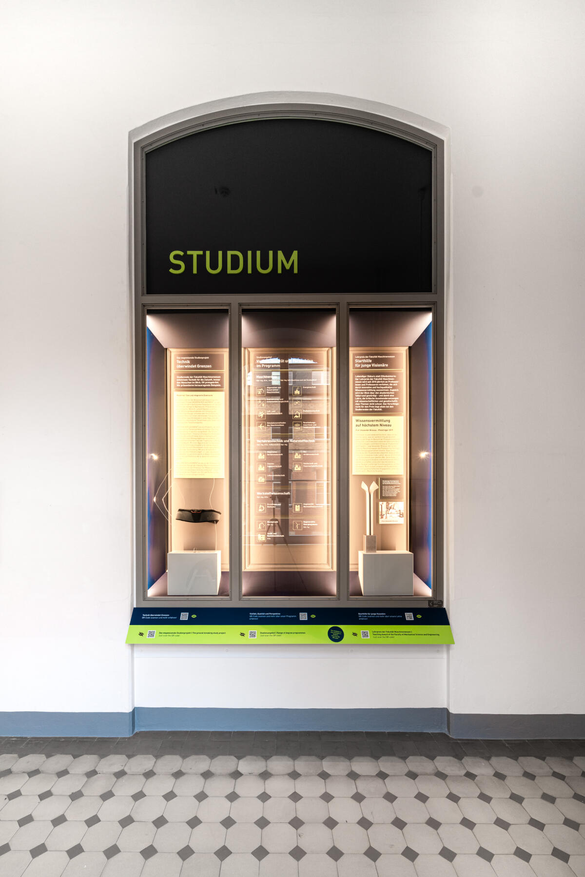 Das Bild zeigt eine große Wandvitrine mit dem Wort 'STUDIUM' in leuchtendem Grün an der Oberseite. In der Vitrine sind mehrere beschriftete Regale und Informationspaneele sichtbar, die zum Thema Studium oder Bildung gehören. Der Boden ist mit einem klassischen, schwarz-weißen Fliesenmuster gestaltet.