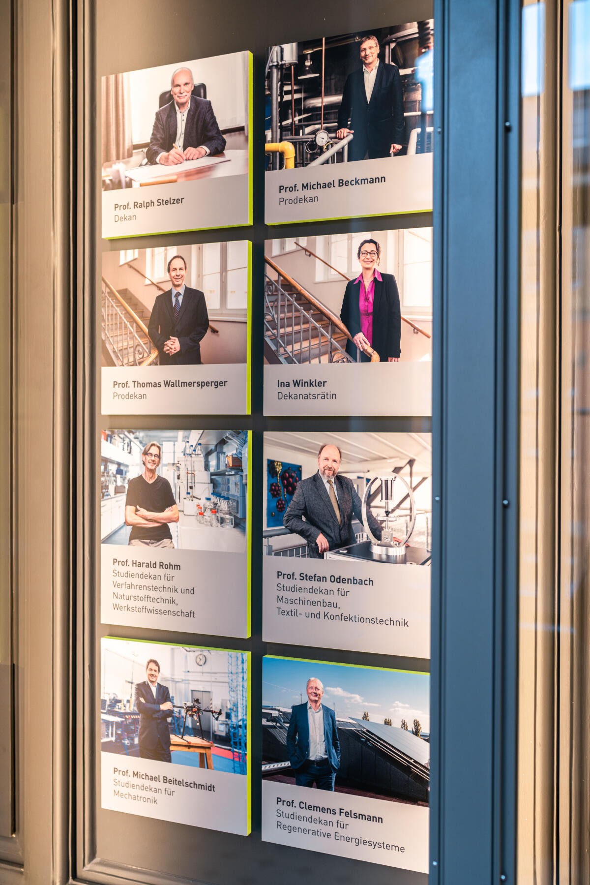 Ein Wanddisplay mit neun Porträts von verschiedenen Personen, die in einem professionellen Umfeld abgebildet sind. Die Bilder sind in drei Reihen und drei Spalten angeordnet, mit Namen und Titeln unter jedem Bild. Die Umgebung wirkt modern und seriös, möglicherweise in einem akademischen oder beruflichen Kontext.