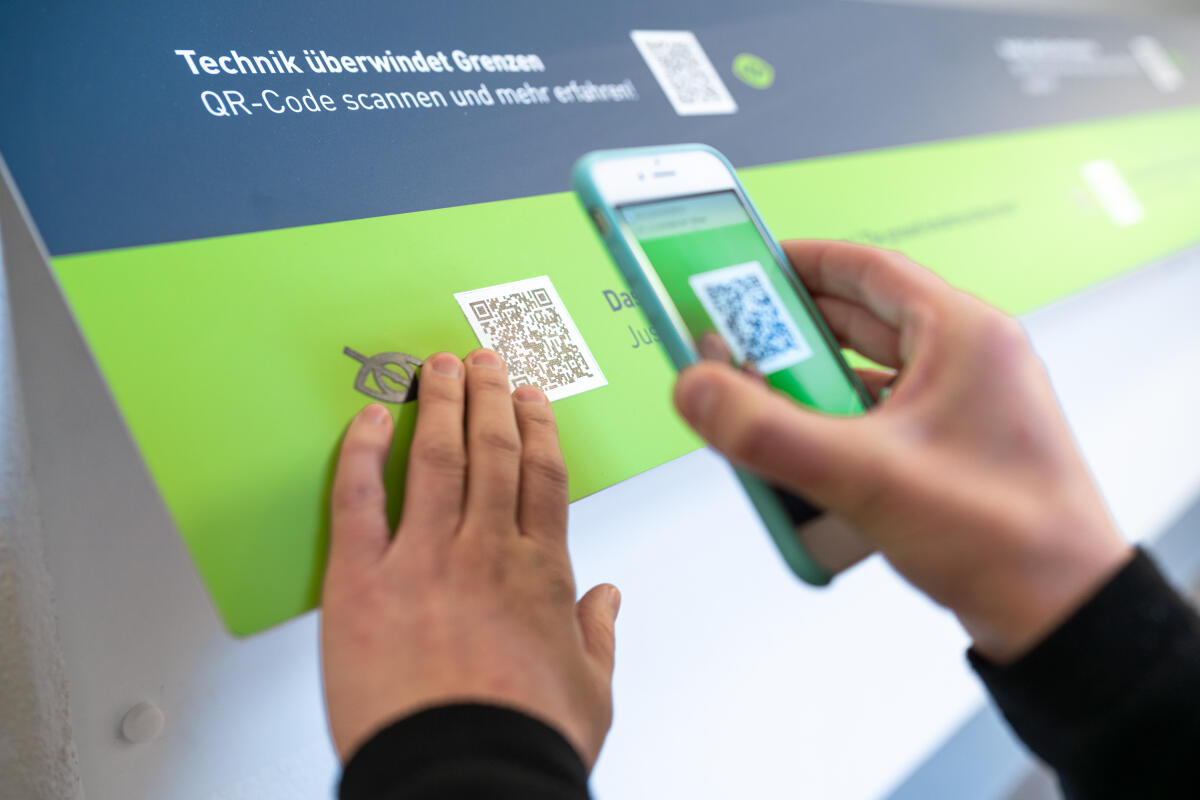 Ein Benutzer scannt mit einem Smartphone einen QR-Code, der auf einem smaragdgrünen Schild angebracht ist. Der Schriftzug auf dem Schild besagt, dass Technik Grenzen überwindet und bietet weitere Informationen an. Die Hand des Benutzers ist auf dem Schild positioniert, während das Smartphone auf den QR-Code gerichtet ist.
