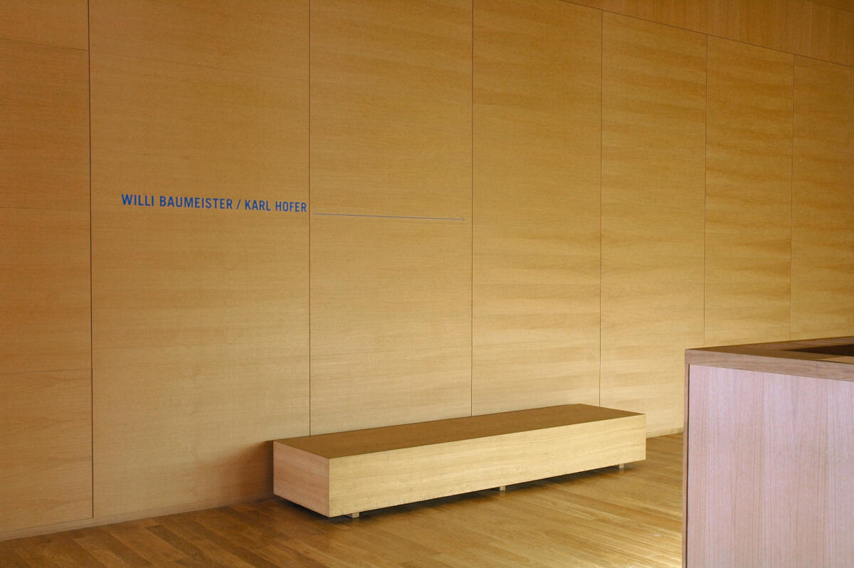 Der Raum hat eine minimalistische Holzverkleidung mit einer langen, schlichten Bank aus Holz an der Wand. An der Wand ist der Schriftzug 'WILLI BAUMSTEIER / KARL HOTEL' in blauer Farbe zu sehen. Der Boden ist aus hellem Holz und verstärkt die ruhige Atmosphäre des Raumes.