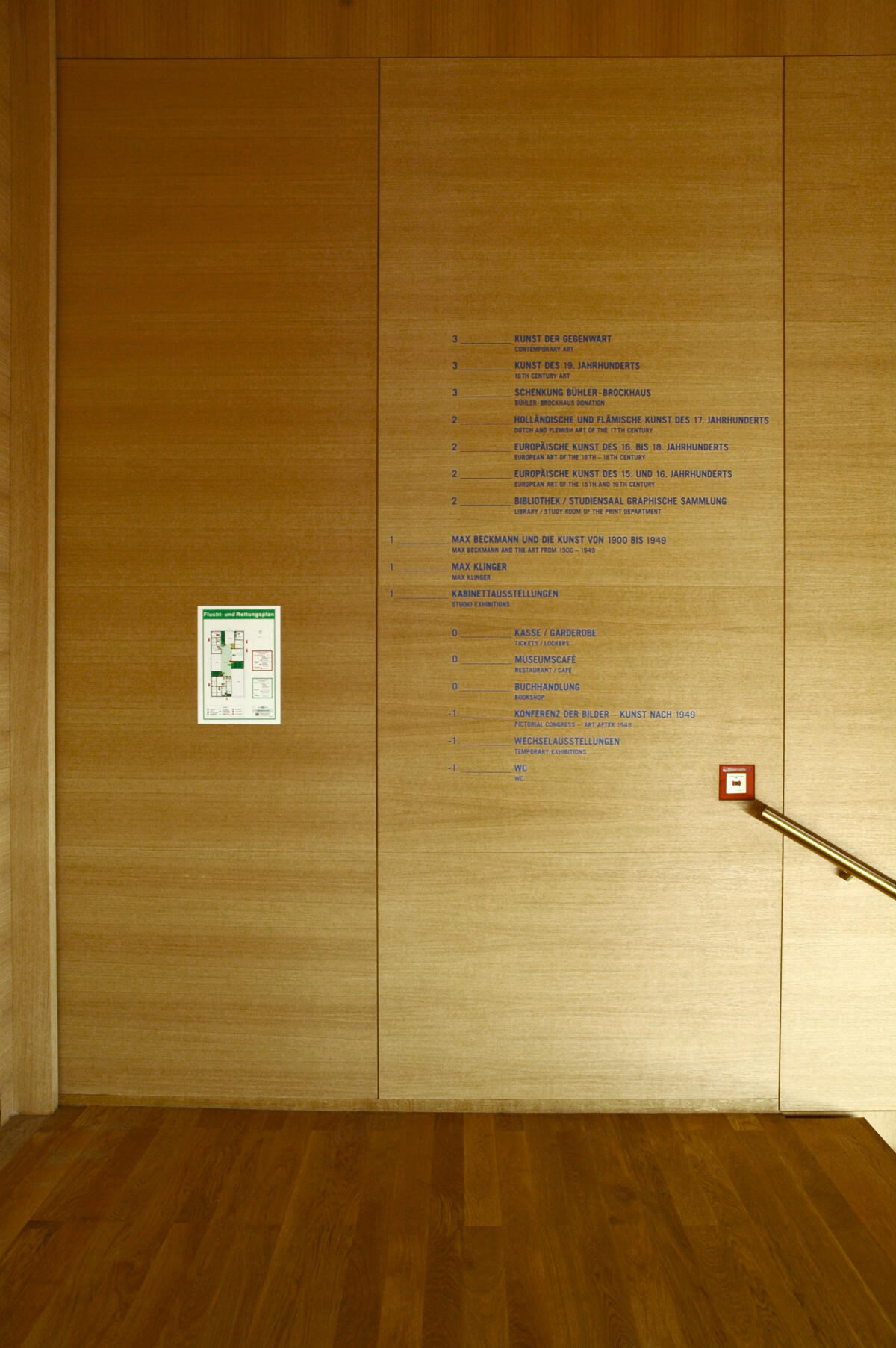 Ein holzgetäfelter Wandraum mit senkrechten Linien, die die Textinformationen strukturieren. Auf der linken Seite befindet sich ein grünes Schild, das einen Notfallplan darstellt, während rechts ein rotes Sicherheitssymbol zu sehen ist. Die Wände sind mit blauen Texten beschriftet, die wichtige Informationen enthalten.