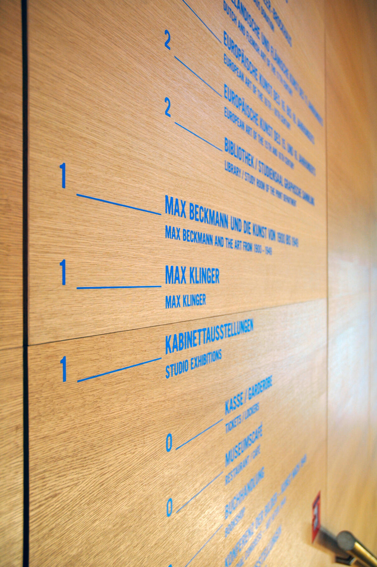 Ein Informationstafeln aus hellem Holz mit blauen Schriftzügen. Die Beschriftung zeigt Namen, möglicherweise von Künstlern oder Räumen, und ist nummeriert. Die Zahlen sind in einer vertikalen Anordnung, was auf eine Art von Wegweiser oder Raumübersicht hinweist.