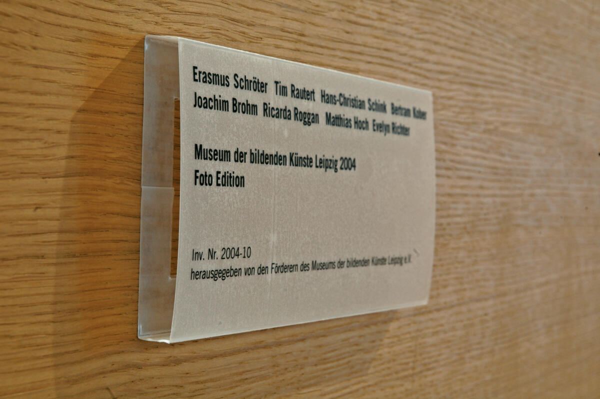 Ein beigefarbenes Schild, das an einer Wand befestigt ist. Es enthält Namen von Künstlern und Informationen über eine Fotoedition aus dem Jahr 2004 des Museums der bildenden Künste in Leipzig. Die Schrift auf dem Schild ist in einer einfachen, klaren Schriftart gehalten.