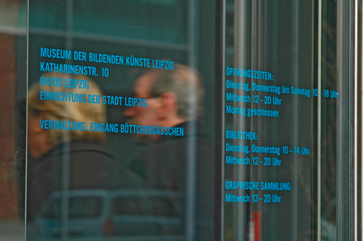 Das Bild zeigt die Glasfassade des Museums der bildenden Künste in Leipzig, auf dem in blauer Schrift der Name des Museums, die Adresse und die Öffnungszeiten abgedruckt sind. Ein Teil von zwei unscharfen Personen ist in der Reflexion der Glasfläche zu sehen. Die Schrift ist klar und modern gestaltet.