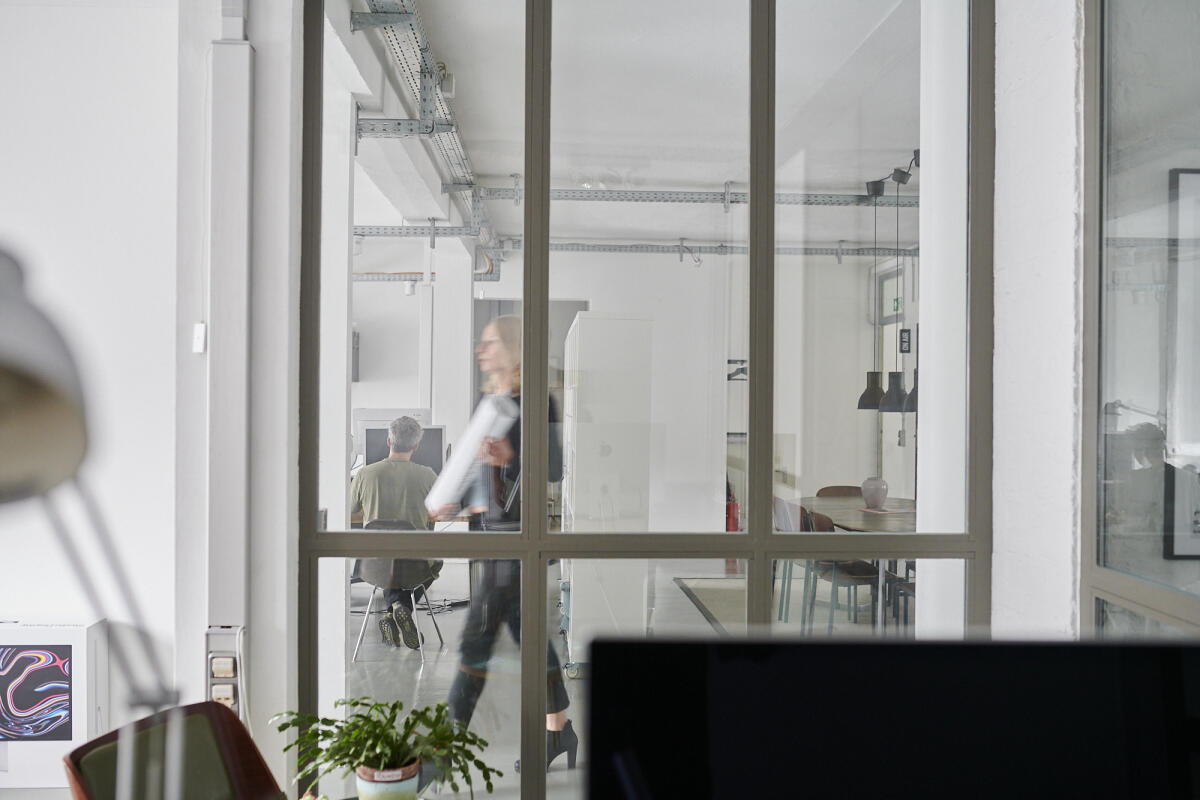 Durch eine gläserne Wand ist ein heller, moderner Büroraum zu sehen. Eine Person mit Dokumenten in der Hand geht vorbei, während im Hintergrund zwei Personen an einem Tisch sitzen. Der Raum ist minimalistisch eingerichtet, und Pflanzen sowie Möbel sind sichtbar.