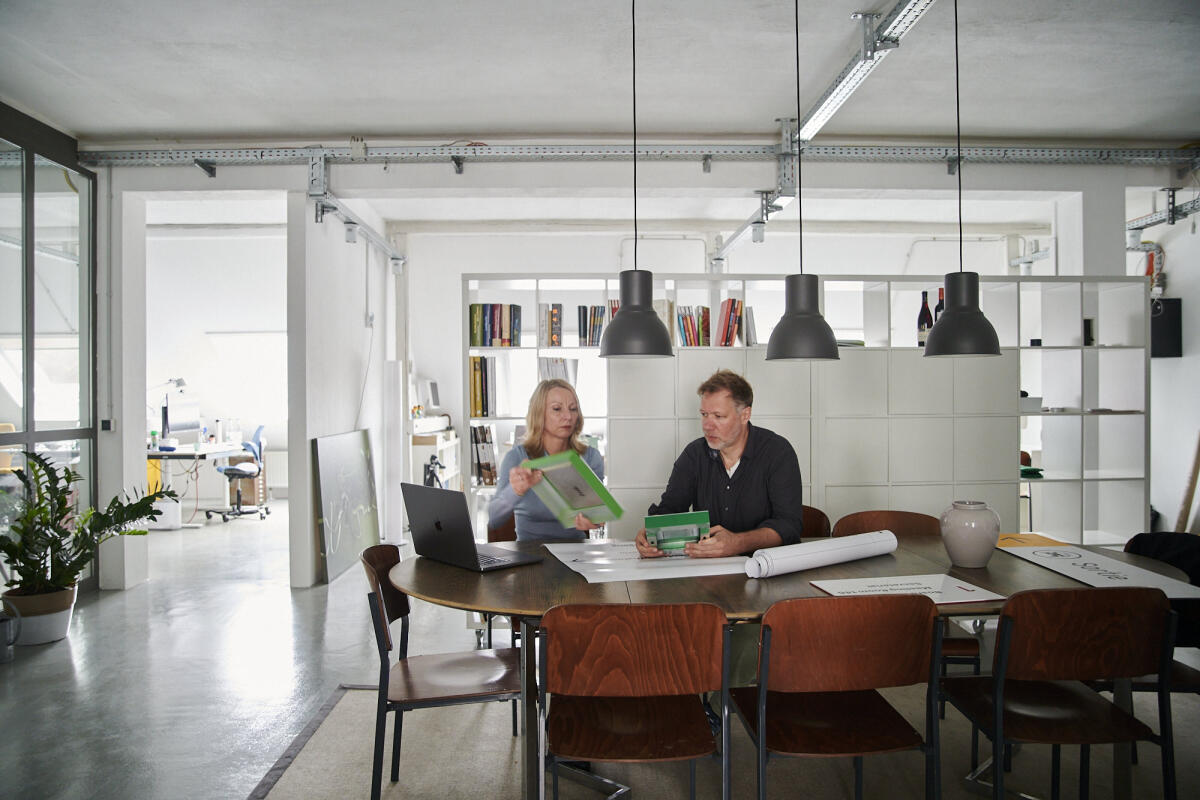 In einem modernen Büroraum sitzen zwei Personen an einem runden Tisch. Eine Frau, die eine grüne Mappe hält, schaut auf einen Mann, der an einem Laptop arbeitet. Im Hintergrund sind Regale mit Büchern und Pflanzen sichtbar, sowie Hängelampen, die den Raum beleuchten.