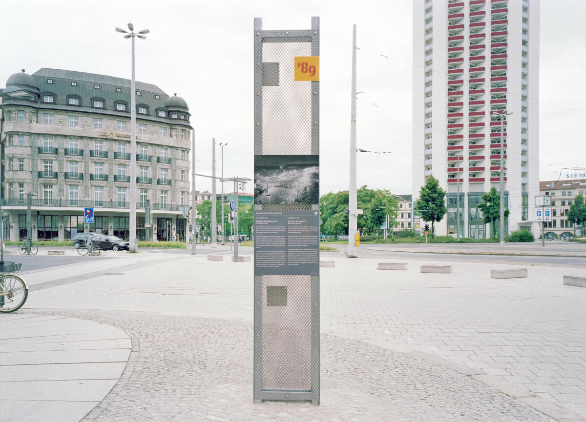 Das Bild zeigt einen hohen, schlanken Informationspylon in einem urbanen Raum, umgeben von einem gepflasterten Platz. Links ist ein mehrstöckiges Gebäude mit einem markanten Dach, während im Hintergrund ein hohes Wohngebäude zu sehen ist. Der Pylon enthält mehrere informative Tafeln, die teilweise mit Bildern und Text versehen sind.