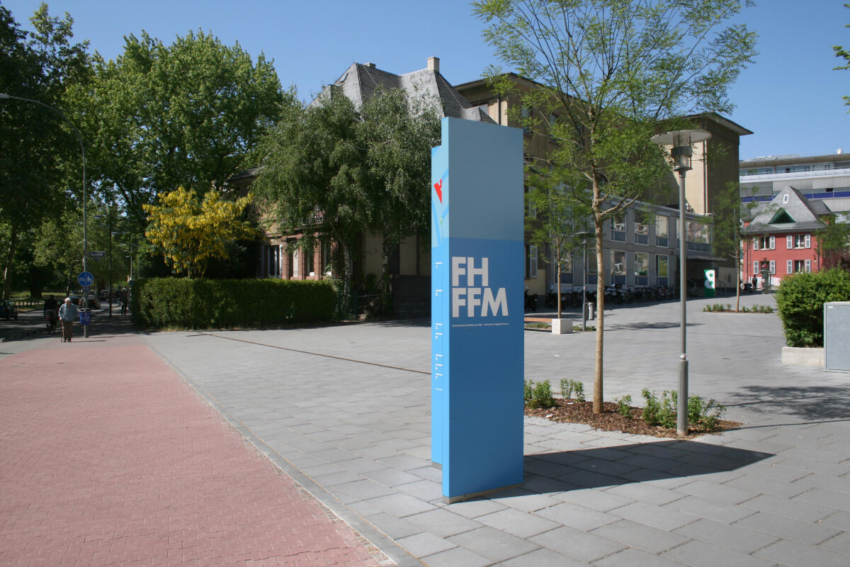 Das Bild zeigt einen modernen, hochstehenden Informationspylon in einem lichtdurchfluteten urbanen Umfeld. Der Pylon ist blau und trägt die Buchstaben 'FH FFM'. Im Hintergrund sind Bäume und ein Gebäude zu sehen, das eine Hochschul- oder Bildungseinrichtung darstellen könnte.