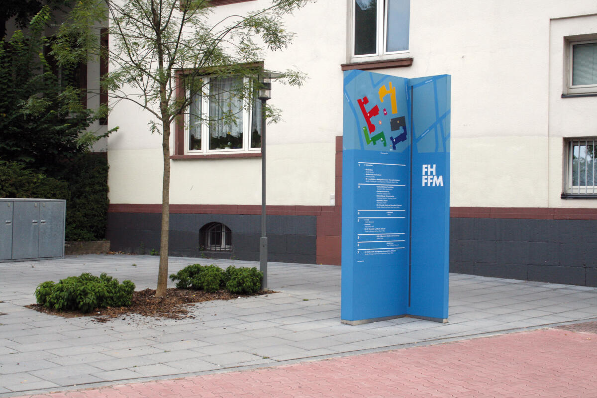 Ein Informationsschild in einem urbanen Bereich, das auf der rechten Seite aufgestellt ist. Es hat eine blaue Farbe mit bunten, stilisierten Buchstaben und einem Logo oben. Der Hintergrund zeigt eine gepflasterte Fläche mit einem jungen Baum und einigen Büschen.