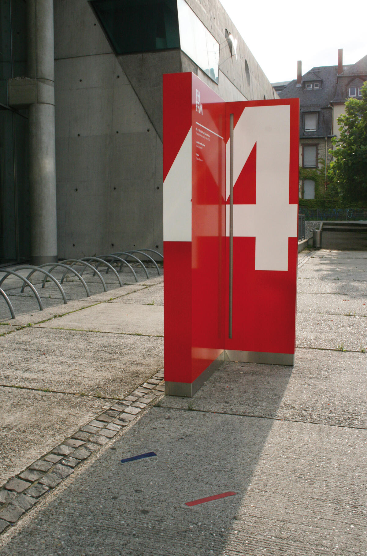 Eine große, rote Stele mit der Zahl 4 in weißer Farbe steht auf einem Platz. Es gibt Betonboden und eine Fahrradabstellanlage.