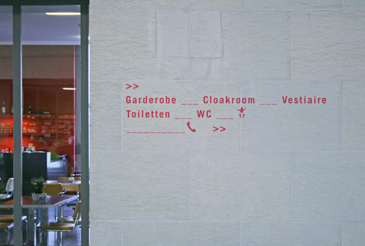 An einer hellen Wand sind rote Orientierungsschilder angebracht, die auf verschiedene Dienstleistungen hinweisen: 'Garderobe', 'Cloakroom', 'Vestiaire' und 'Toiletten'. Eine Telefon-Symbol und Pfeile deuten zusätzlich den Weg an. Im Hintergrund sind unscharf Tische und Stühle zu erkennen, die Teil eines Cafés sein könnten.