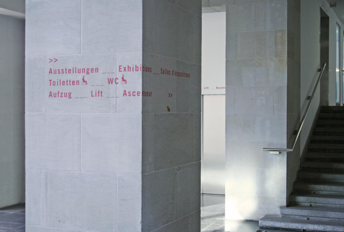 Ein steinerner Säulenpfeiler in einem Innenraum, der mit roten Schriftzeichen beschriftet ist. Die Beschriftungen weisen auf Ausstellungen, Toiletten, Aufzüge und Treppen hin. Im Hintergrund sind Treppen und ein Aufzug sichtbar, die den Weg in verschiedene Richtungen zeigen.