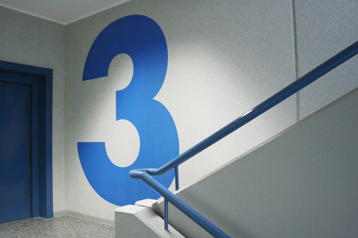 Ein Treppenhaus mit einem großen, blauen Ziffernzeichen '3' an der Wand. Links befindet sich eine blaue Tür, die den Zugang zu einem anderen Bereich markiert. Eine graue Treppe führt nach oben, und ein blauer Handlauf verläuft entlang der Treppe.