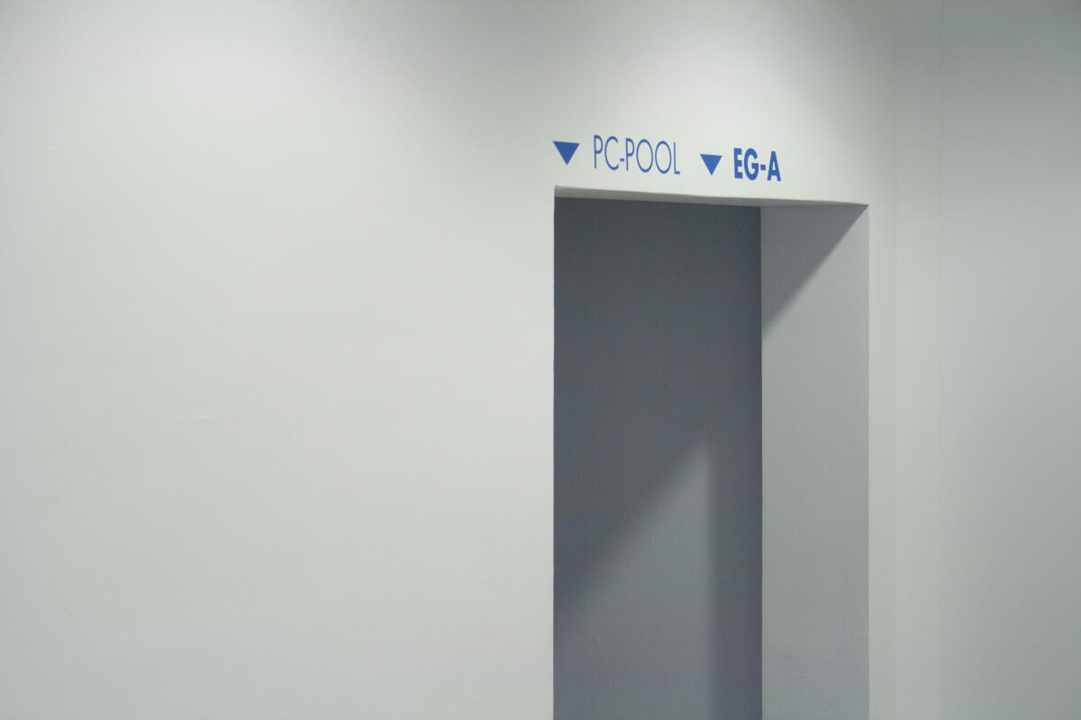 Ein schlichter, weißer Wandraum mit einer offenen Tür. Über der Tür sind zwei Begriffe in blauer Schrift angebracht: 'PC-POOL' und 'EG-A', wobei ein Pfeil nach unten zeigt. Die Wand selbst ist hell und reflektiert das Licht.